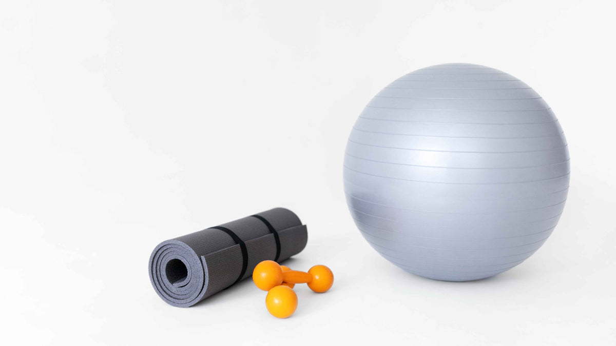 Fitness-Set mit grauer Gymnastikball, schwarzer Yogamatte und orangefarbenen Hanteln auf weißem Grund.