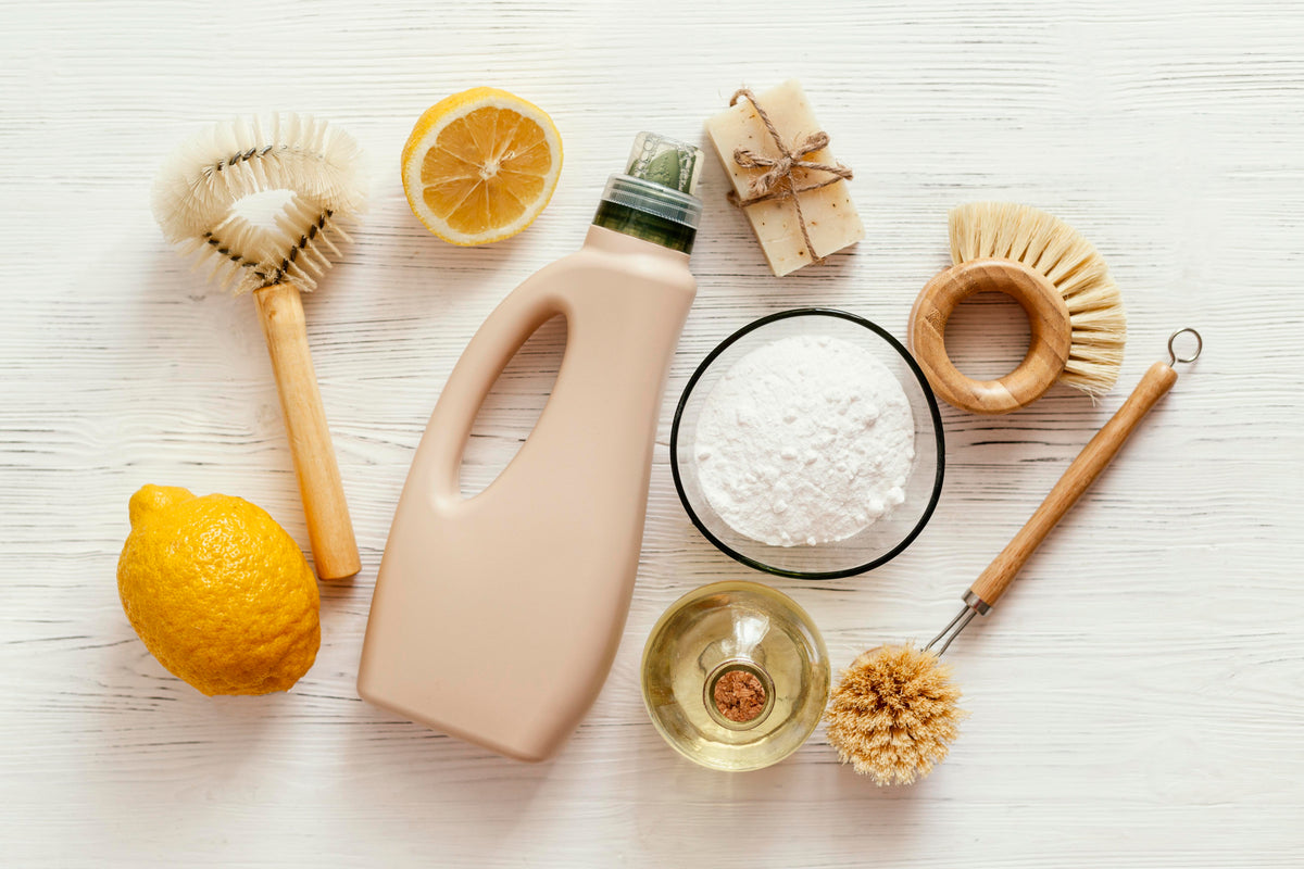 Nachhaltige Reinigungsmittel: Bio-Spülmittel, Natron, Zitrone und Holzbürsten auf weißem Holz.