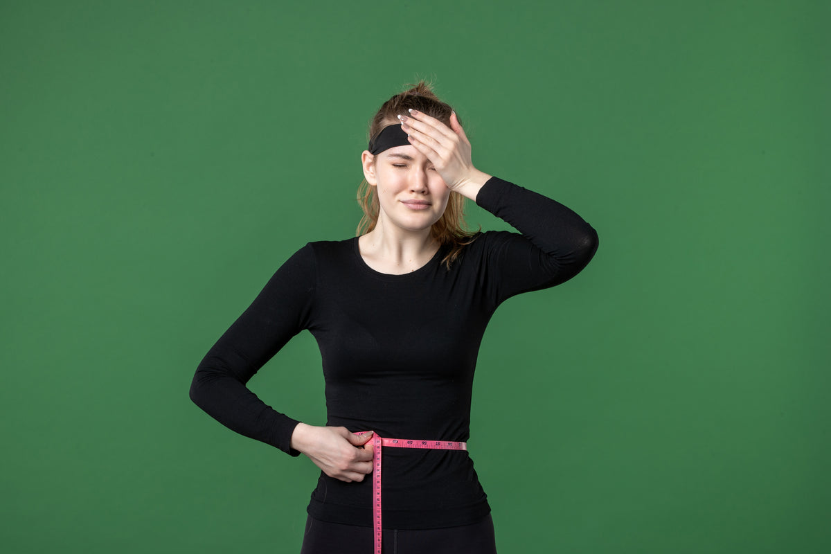 Frustrierte Frau in Sportkleidung mit Maßband um die Taille vor grünem Hintergrund.