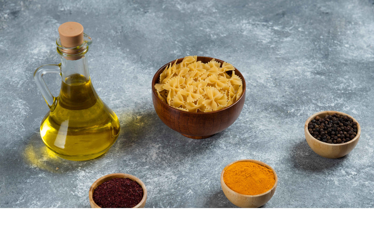 Olivenöl in Glasflasche, Farfalle-Pasta in Holzschale und verschiedene Gewürze auf grauem Untergrund.