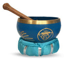 Eine blaue, goldverzierte Klangschale mit dem Vishuddha-Chakra-Symbol und Klöppel liegt auf einem türkisfarbenen Kissen.