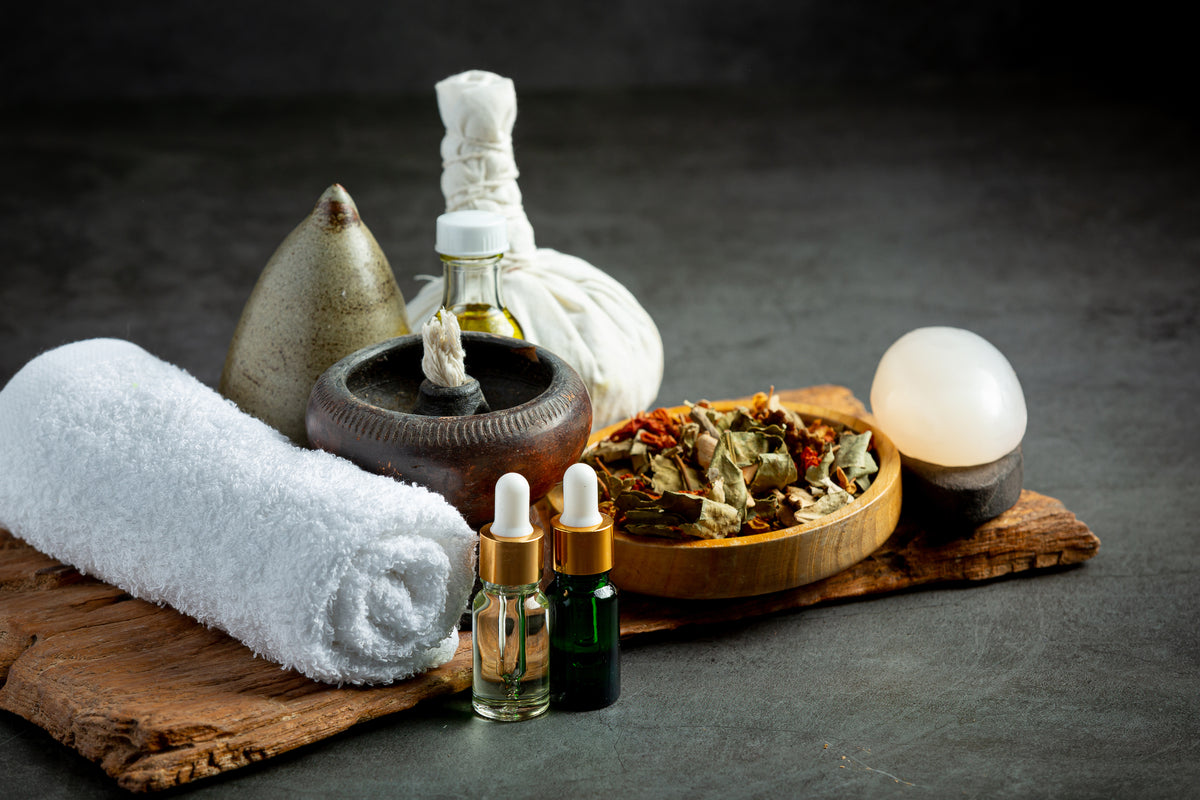 Wellness-Set mit Kräuterbeuteln, ätherischen Ölen, weißem Handtuch und getrockneten Kräutern.