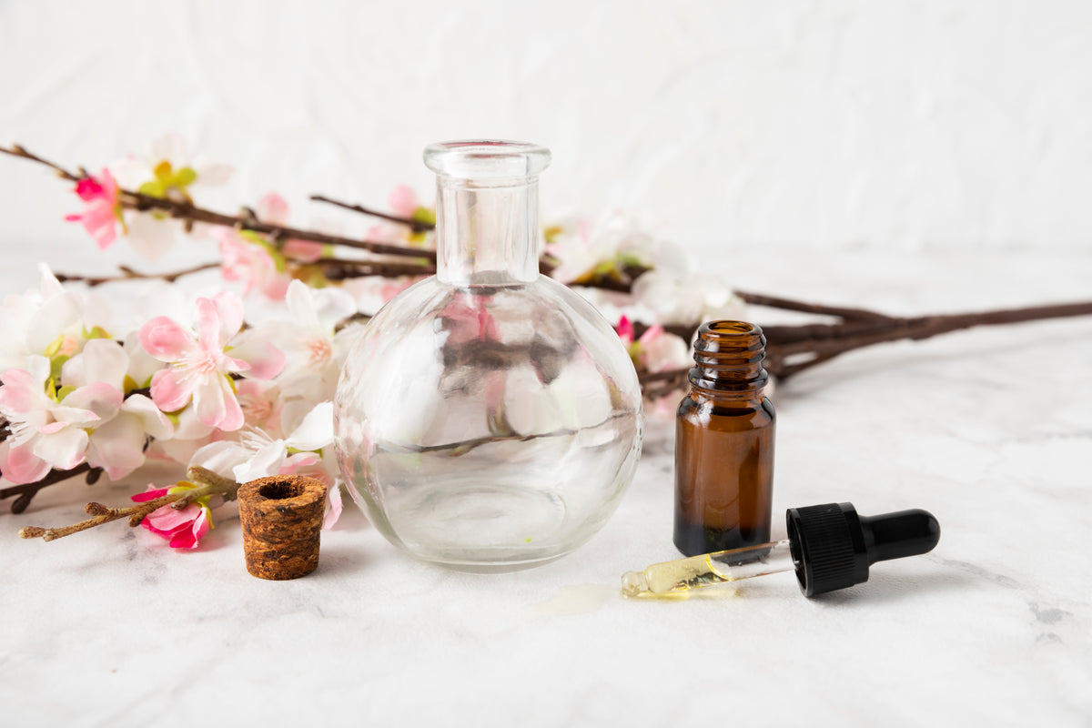 Braune Glasflasche mit Pipette und leere Glasvase vor Kirschblüten auf Marmor.