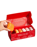 Eine hand hält ein orange transparentes ovales kojisäure-hautaufhellendes koj white soap über einer roten box mit sechs pastellfarbenen koj white gift bars