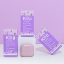 Lilafarbenes Bild mit zwei Schachteln Koji White Brightening Soap mit Kojisäure und Hyaluronsäure. Davor befindet sich ein rosa Seifenstück.