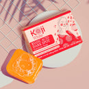 Ein Stück orangefarbene Kojic Acid Seife neben der rosa-weißen Verpackung.
