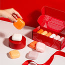 Eine Hand hält ein orangefarbenes Seifenstück über einer Sammlung bunter Seifen und einer offenen roten Geschenkbox.