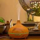 Ein tropfenförmiger Aroma-Diffuser in Holzoptik, der einen feinen Nebel ausstößt und von einem grünen LED-Ring beleuchtet wird, platziert in einem gemütlich beleuchteten Raum.