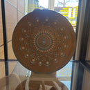 Eine goldfarben lackierte Vinyl-Schallplatte mit einem kunstvollen Mandala aus weißen und türkisfarbenen Punkten, aufgestellt auf einem Ständer vor einem Fenster.