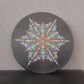 Eine schwarze Vinyl-Schallplatte, verziert mit einem pastellfarbenen Punktmalerei-Mandala in symmetrischer Sternform und kleinen Strasssteinen, angelehnt an eine graue Wand.