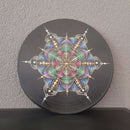 Eine schwarze Vinyl-Schallplatte, verziert mit einem pastellfarbenen Punktmalerei-Mandala in symmetrischer Sternform und kleinen Strasssteinen, angelehnt an eine graue Wand.