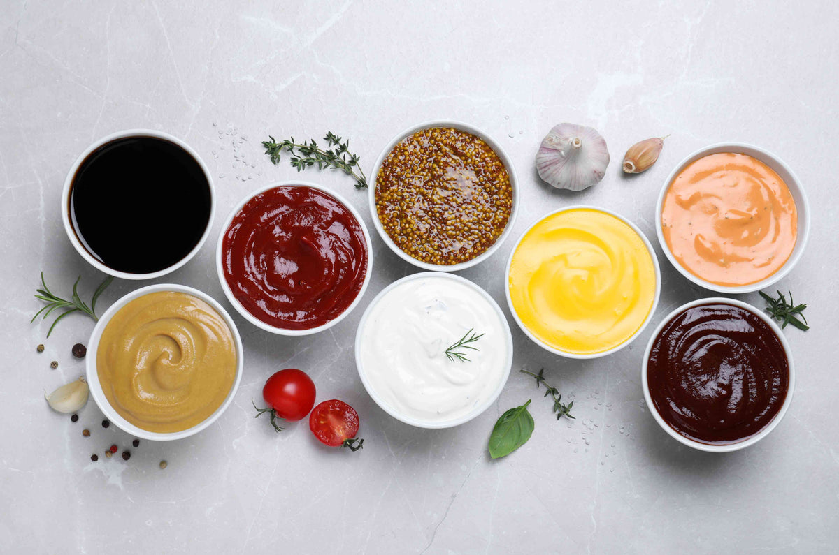 Verschiedene Saucen und Dips in weißen Schalen mit Kräutern und Tomaten auf hellem Untergrund.