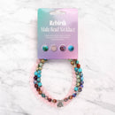 Eine verpackte Myga Rebirth Mala-Perlenkette mit bunten Edelsteinen, einer Kette aus rosa Quarz und einem silbernen Lotus-Anhänger auf einer Marmoroberfläche.