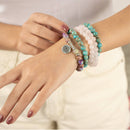 Eine Person, die ein mehrreihiges Perlenarmband mit rosa, türkisfarbenen und lila Steinen und einem silbernen Lotus-Anhänger trägt.