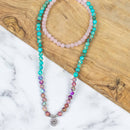Eine lange Mala-Perlenkette mit Türkis-, Rosenquarz- und lila Steinen sowie einem silbernen Lotus-Anhänger auf einem Untergrund aus Marmor und Holz.