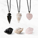 Drei Kristallanhänger an schwarzen Kordeln (schwarze Obsidian-Pfeilspitze, klare Quarz-Geoden-Pfeilspitze, herzförmiger rosa Rosenquarz) liegen über den entsprechenden rohen Kristallen auf weißem Marmoruntergrund.