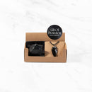 Eine kleine braune Papp-Geschenkbox mit einem rohen Stück schwarzem Obsidian und einem passenden Obsidian-Pfeilspitzen-Anhänger an einer schwarzen Kordel, beschriftet mit „Gift of Protection“ (Geschenk des Schutzes).