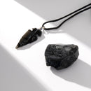 Ein Stück roher schwarzer Obsidian liegt neben einem passenden Obsidian-Pfeilspitzen-Anhänger an einer schwarzen Kordel, auf einer weißen Oberfläche mit starker gerichteter Beleuchtung fotografiert.