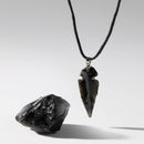 Ein Stück roher schwarzer Obsidian liegt neben einem hängenden Obsidian-Pfeilspitzen-Anhänger an einer schwarzen Kordel, auf weißem Hintergrund mit dramatischen Schatten fotografiert.