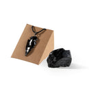 Ein glänzender, geschnitzter schwarzer Obsidian-Pfeilspitzen-Anhänger an einer schwarzen Kordel, präsentiert auf einem kleinen Schmuckständer aus Pappe vor weißem Marmorhintergrund.
