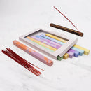 Ein Box-Set mit Räucherstäbchen zum Thema Chakren in pastellfarbenen Schachteln, eine separate Schachtel mit roten Wurzel-Chakra-Räucherstäbchen und ein hölzerner Räucherstäbchenhalter mit einem brennenden Stab auf einer Marmoroberfläche.