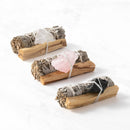 Drei Bündel aus weißem Salbei und Palo-Santo-Holz sind mit Rohkristallen (Bergkristall, Rosenquarz und schwarzer Turmalin/Schungit) und Schnur zusammengebunden und liegen auf einer weißen Marmoroberfläche.