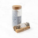 Ein „Clarity“ Salbei-Ritual-Set in einem hohen Glasgefäß mit Korkdeckel, das weißen Salbei und Pulver enthält, steht neben einem Bündel aus weißem Salbei, Palo-Santo-Holz und Bergkristall, alles auf einer weißen Marmoroberfläche.