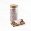 Ein „Love“ Salbei-Ritual-Set in einem hohen Glasgefäß mit Korkdeckel, das weißen Salbei und Pulver enthält, steht neben einem Bündel aus weißem Salbei, Palo-Santo-Holz und einem Rosenquarz-Rohstein, alles auf einer weißen Marmoroberfläche.