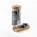 Ein „Protection“ Salbei-Ritual-Set in einem hohen Glasgefäß mit Korkdeckel steht neben einem Bündel aus weißem Salbei, Palo-Santo-Holz und einem schwarzen Turmalin/Schungit-Kristall, alles auf einer weißen Marmoroberfläche.