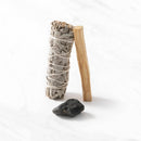 Ein Räucherset, bestehend aus einem Bündel weißem Salbei, einem Stock Palo-Santo-Holz und einem rohen schwarzen Stein, die auf einer weißen Marmoroberfläche aufrecht stehen.