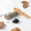 Ein Bündel weißer Salbei und ein Stock Palo Santo Holz liegen auf einer weißen Marmoroberfläche neben einem schwarzen Stein und getrockneten Blättern.