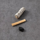 Ein Bündel weißer Salbei und ein Stück Palo Santo Holz liegen zusammen mit einem kleinen schwarzen Stein auf einem dunkelgrauen Hintergrund.