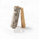 Ein Bündel weißer Salbei und ein Stück Palo Santo Holz stehen aufrecht und lehnen aneinander, mit einem klaren Quarzkristall davor auf einer weißen Marmoroberfläche.