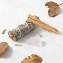 Ein Bündel weißer Salbei und ein Stück Palo Santo Holz liegen auf einer weißen Marmoroberfläche mit einem klaren Quarzkristall und verstreuten, getrockneten Blättern.