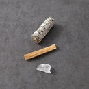 Ein Bündel weißer Salbei und ein Stück Palo Santo Holz liegen zusammen mit einem kleinen klaren Quarzkristall auf einem dunkelgrauen Hintergrund.