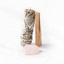 Ein Bündel weißer Salbei und ein Stück Palo Santo Holz stehen aufrecht auf einer weißen Marmoroberfläche, mit einem Rosenquarzkristall davor.