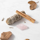 Ein Bündel weißer Salbei und ein Stück Palo Santo Holz liegen auf einer weißen Marmoroberfläche mit einem Rosenquarzkristall und verstreuten, getrockneten Blättern.