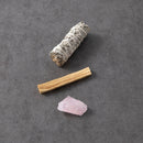 Ein Bündel weißer Salbei und ein Stück Palo Santo Holz liegen zusammen mit einem kleinen Rosenquarzkristall auf einem dunkelgrauen Hintergrund.