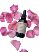 Ein dunkelbernsteinfarbener glas-sprayflakon mit schwarzer düse und paavani ayurveda rose water etikett umgeben von rosa rosenblättern für organisches bulgarisches beruhigendes aromatherapie-nebel