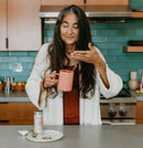 Eine Frau mit langen, grau melierten Haaren steht in einer Küche, hält eine Tasse Tee und riecht daran; eine Dose Woman's Blend Tee steht auf der Theke.