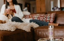 Eine Dose Paavani Ayurveda Woman's Blend Tee steht im Vordergrund auf einem Tisch, im Hintergrund entspannt eine Frau auf einem braunen Ledersofa.