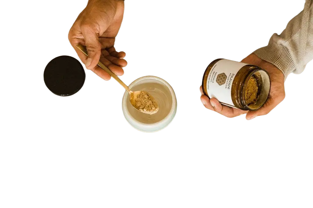 Ein glas mit paavani ayurveda triphala pulver in brauner farbe und weißem etikett wird in der hand gehalten und in eine schale gegeben