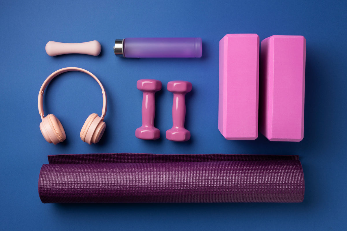 Yoga-Zubehör Set mit lila Matte, Hanteln, Blöcken, Trinkflasche und Kopfhörern auf blauem Grund.
