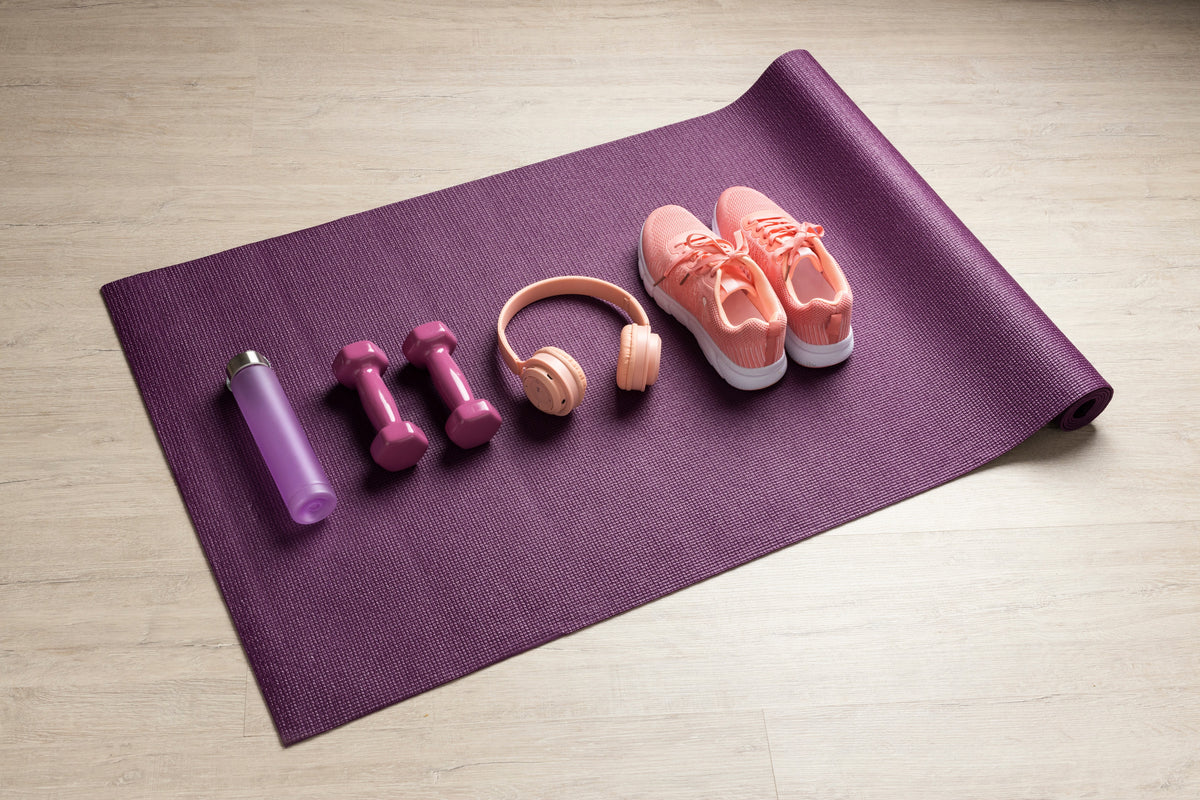 Lila Yogamatte mit pinken Hanteln, Kopfhörern, Sportschuhen und Wasserflasche auf Holzgrund.