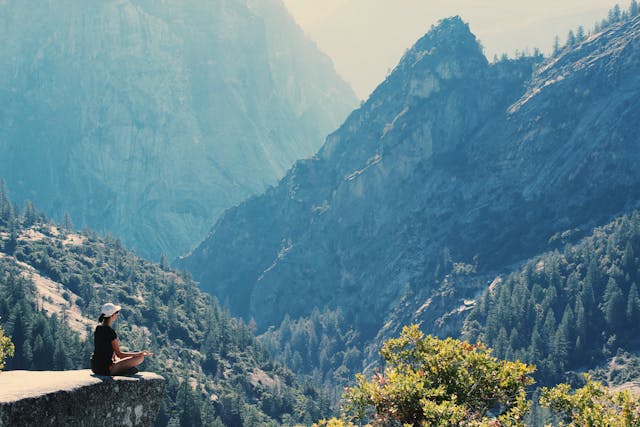 Eine Person meditiert auf einer Klippe mit Blick auf ein weites Bergtal.