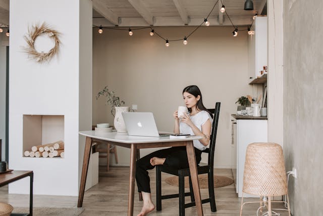 Eine Frau, die mit einem Laptop an einem Esstisch sitzt und in einem gemütlichen Wohninterieur eine Tasse hält.