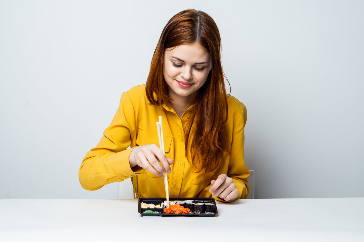 Junge Frau mit rötlichen Haaren in gelber Bluse isst Sushi mit Stäbchen.