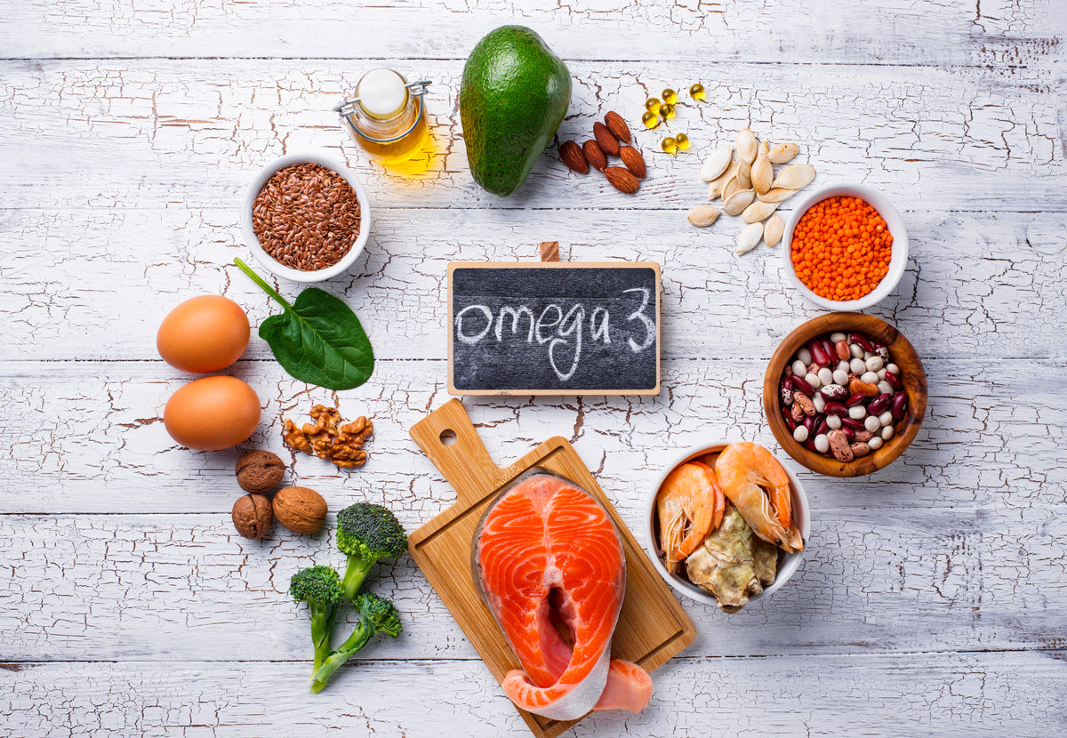 Gesunde Omega-3 Lebensmittel wie Lachs, Eier, Avocado, Nüsse und Samen auf Holzgrund.
