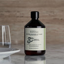 Eine Lifestyle-Aufnahme einer braunen Flasche „Read/the/label“ Spülmittel mit Bergamotte & Grosso-Lavendel auf einer Marmoroberfläche neben einem Glas Wasser und einem weißen Teller.