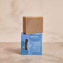 Apeiron Fuß- & Hornhaut Peeling-Seife und ihre blaue Verpackung auf einer Steinfläche mit sanften Pflanzenschatten.
